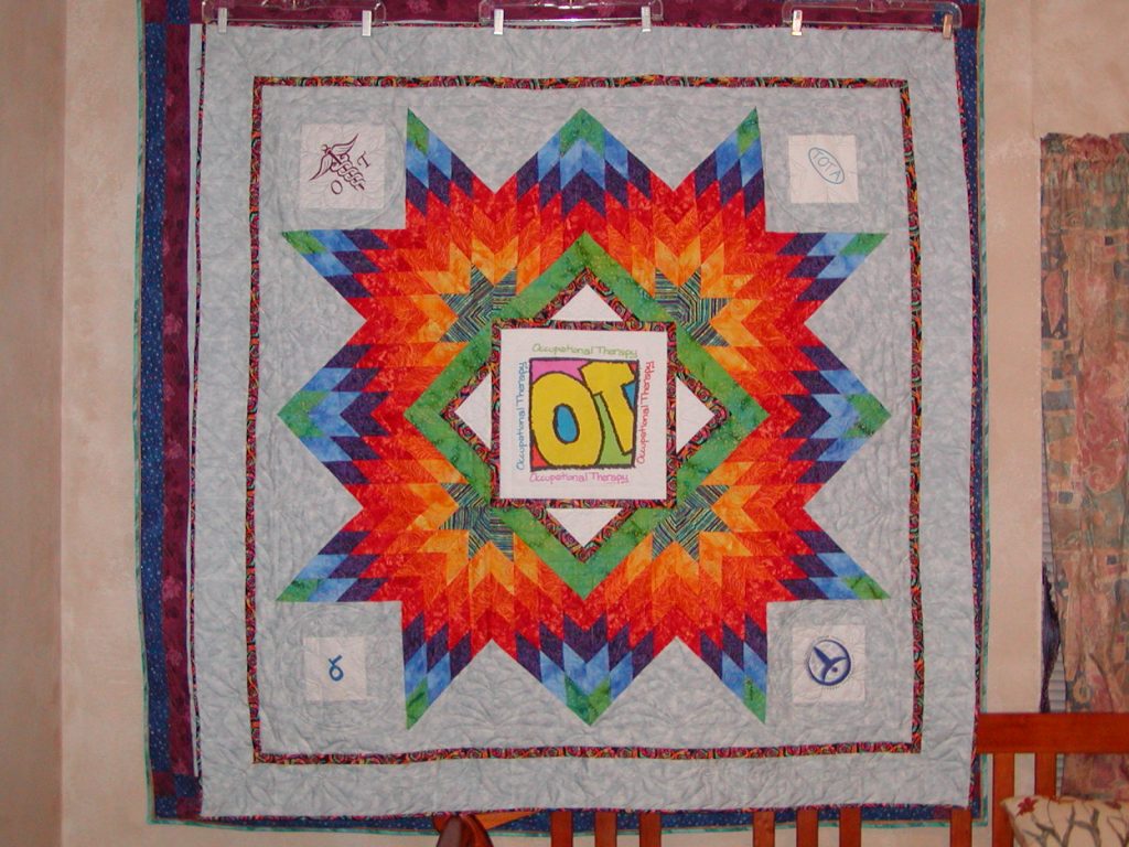 A colorful quilt featuring a star design with various geometric patterns and vibrant colors, centered around the text 'Occupational Therapy'. The quilt is displayed on a wall with decorative elements around it.